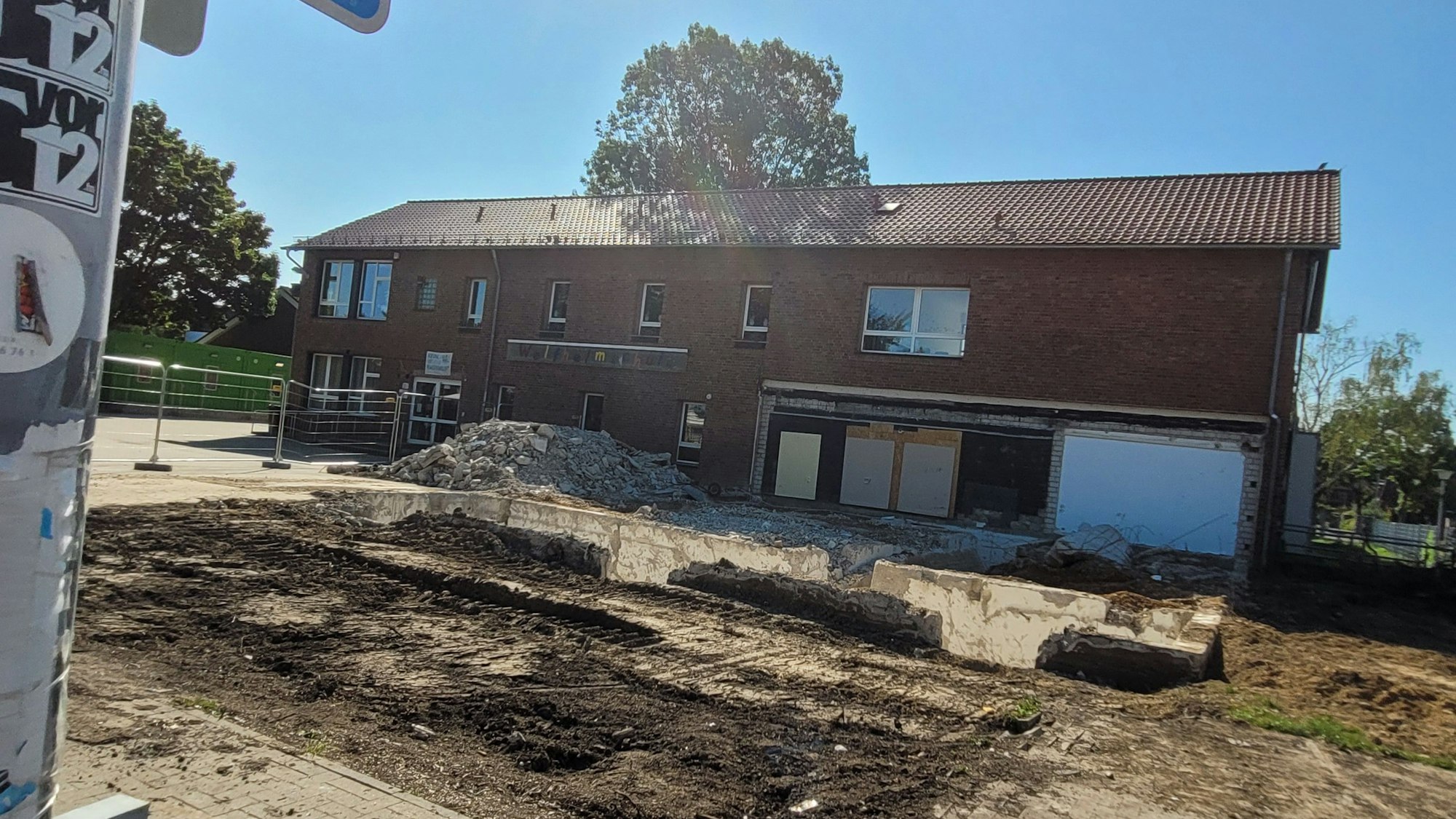 Im Hintergrund ein Gebäude mit Klinkerfassade, davor eine freie Fläche, in der sich noch Mauerreste befinden.