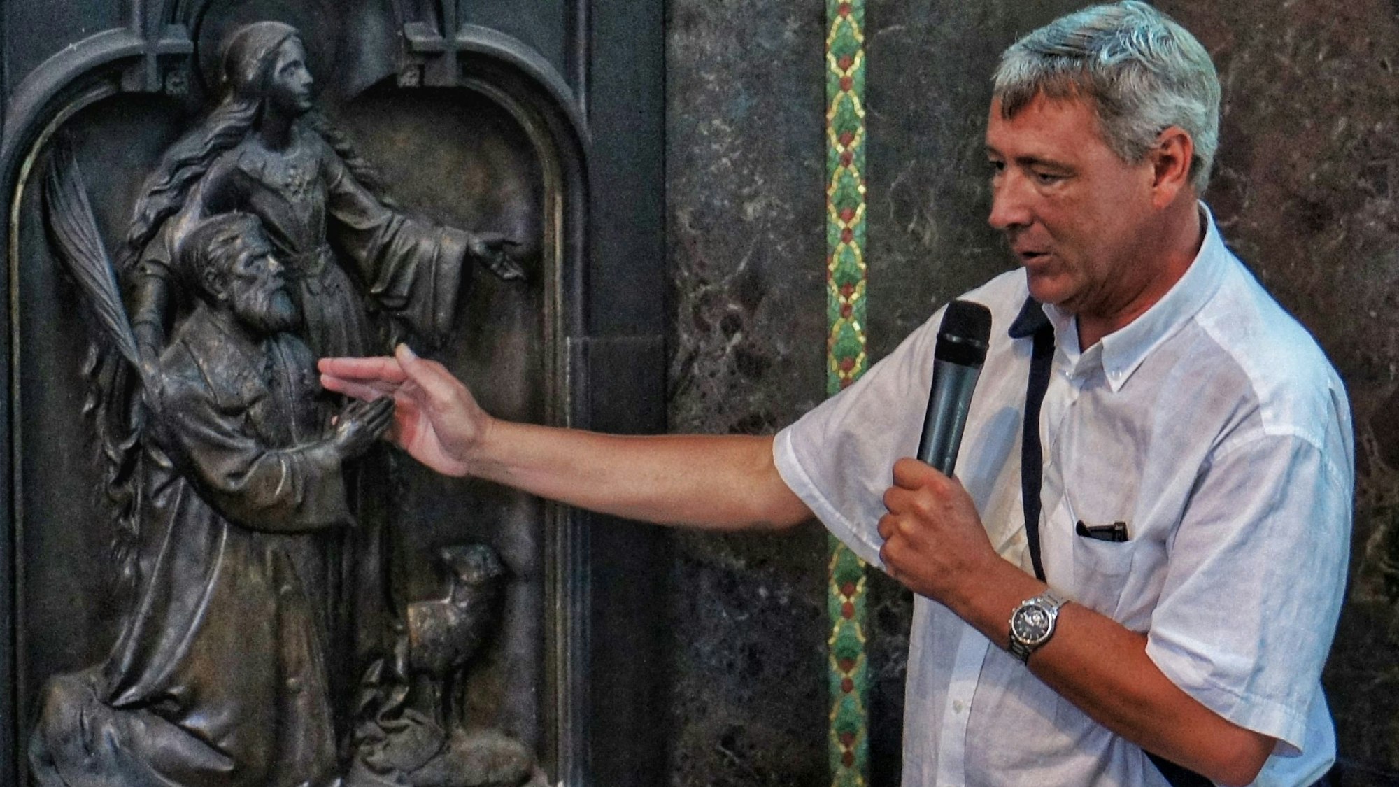 Pfarrer Frank Müller (1962 bis 2025) zeigt ein Bronzerelief in der Kölner Kirche Sankt Agnes mit dem Stifter der Kirche, Peter Joseph Roeckerath.