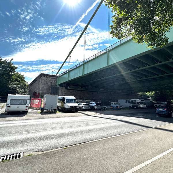 Unter einer großen Straßenbrücke verläuft eine mehrspurige Straße, neben der Straße parken einige Autos und Campingwagen unter der Brücke.