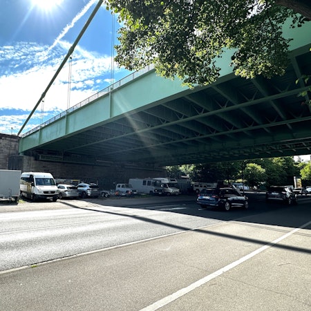 Unter einer großen Straßenbrücke verläuft eine mehrspurige Straße, neben der Straße parken einige Autos und Campingwagen unter der Brücke.
