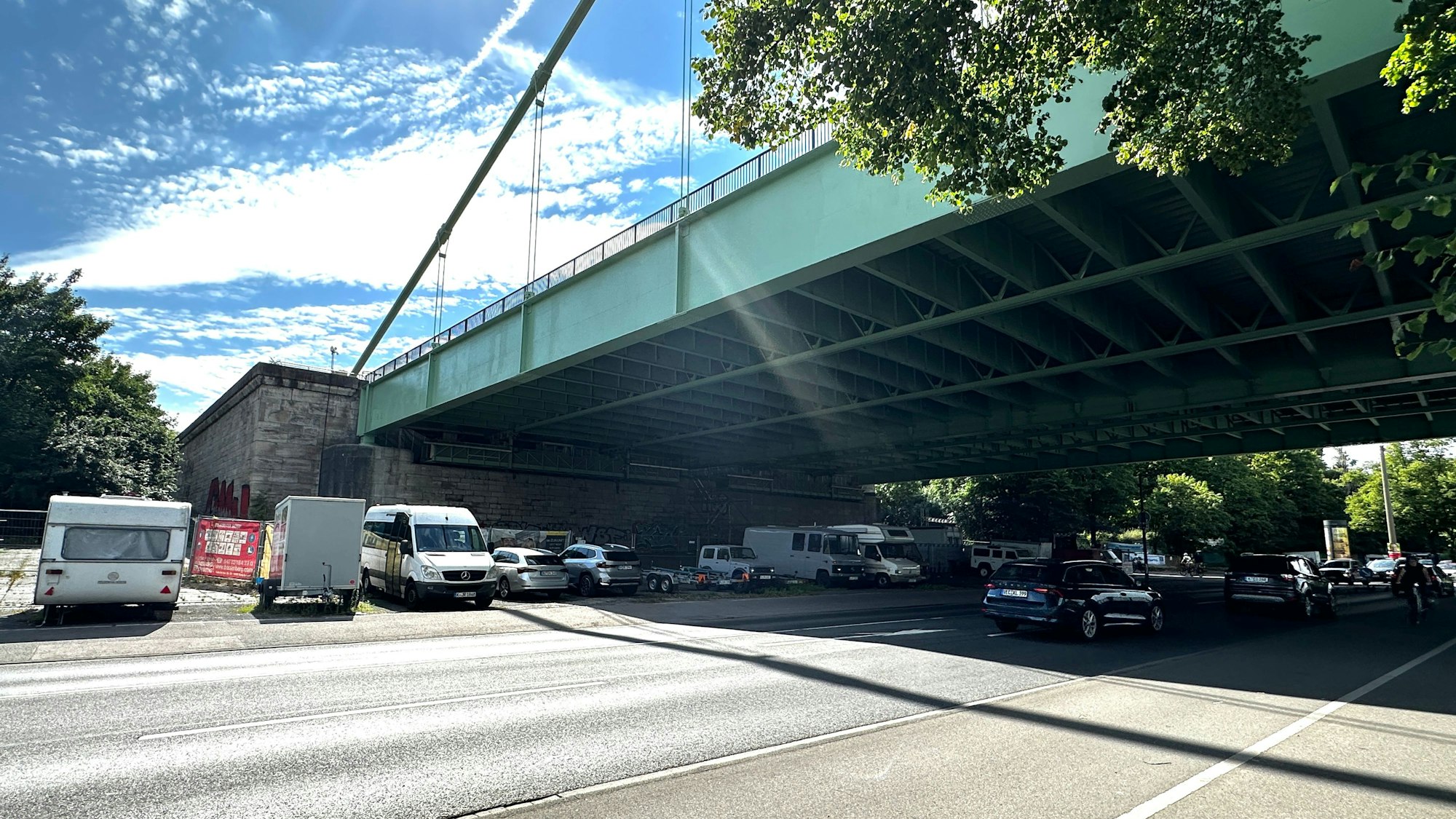 Unter einer großen Straßenbrücke verläuft eine mehrspurige Straße, neben der Straße parken einige Autos und Campingwagen unter der Brücke.