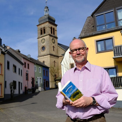 Der Schriftsteller Ralf Kramp hält sein Buch ‚Gebrauchsanweisung für die Eifel‘ in der Hand. Er steht vor einem Altstadt-Ensemble mit bunt angestrichenen Häusern in Hillesheim, imm Hintergrund ragt der Turm der Pfarrkirche St. Martin empor.