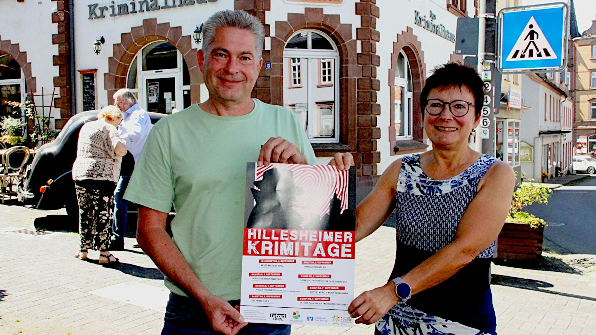 Frank Reuter und Rita Kallenberg stehen vor dem „Kriminalhaus“ und halten ein Plakat der Krimitage in den Händen.