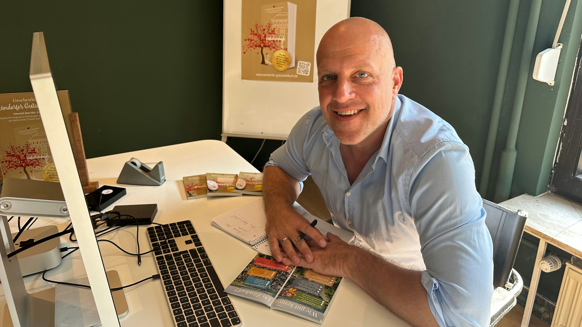 Ein Mann mit Glatze, der ein hellblaues Hemd trägt, sitzt an einem Schreibtisch.