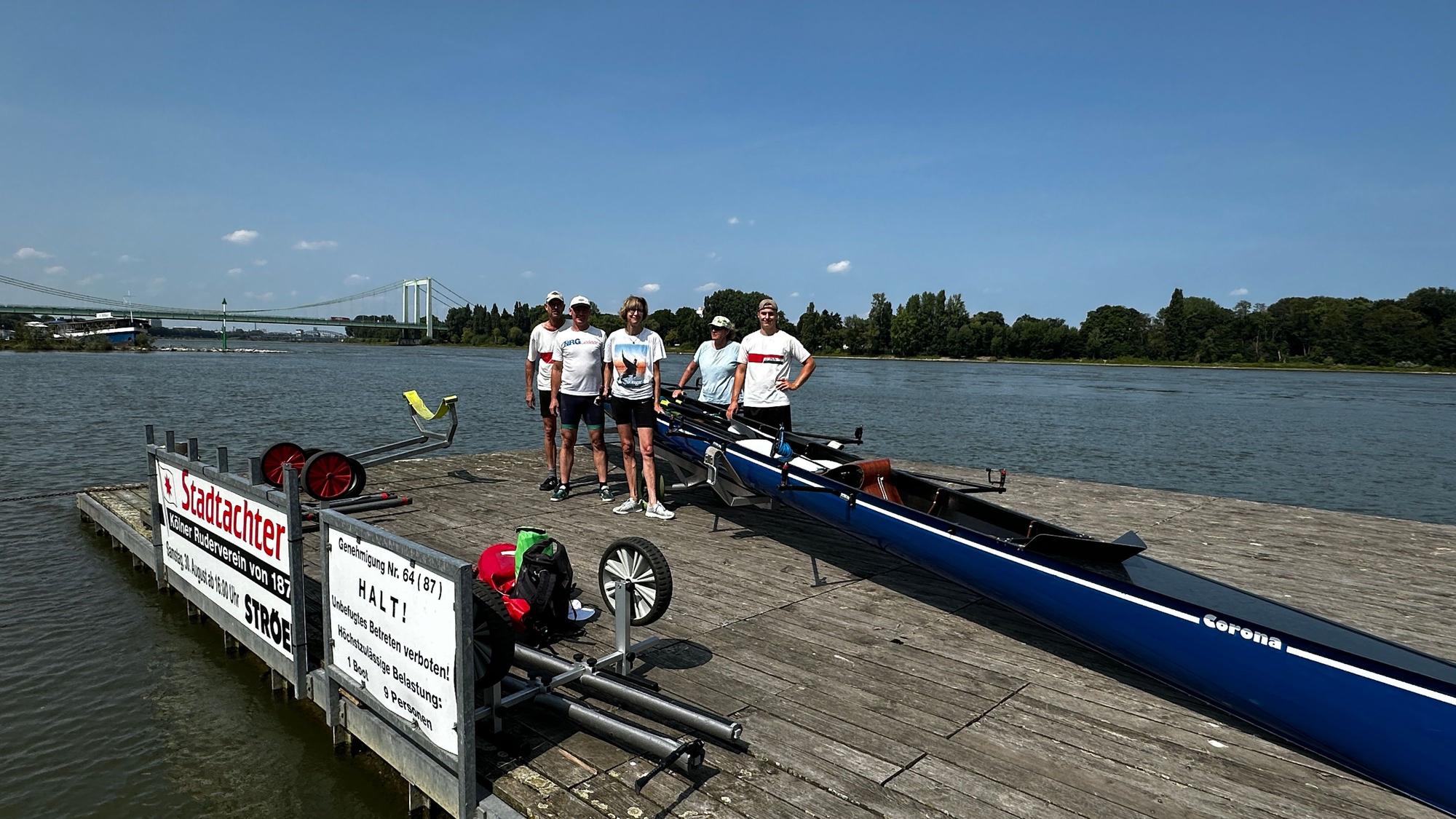 Auf einem Bootssteg stehen fünf Menschen neben einem Ruderboot, im Hintergrund sind der Rhein und die Rodenkirchener Brücke zu sehen.
