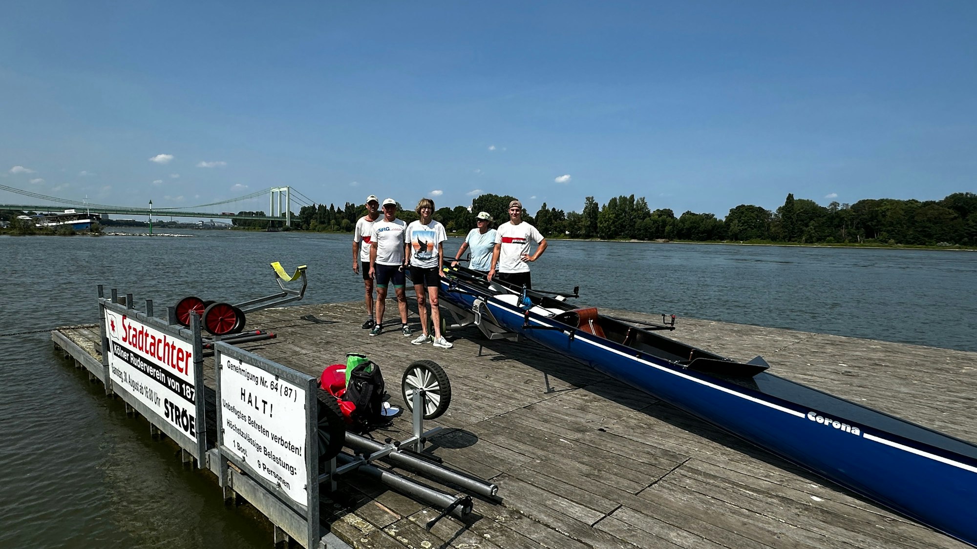 Auf einem Bootssteg stehen fünf Menschen neben einem Ruderboot, im Hintergrund sind der Rhein und die Rodenkirchener Brücke zu sehen.
