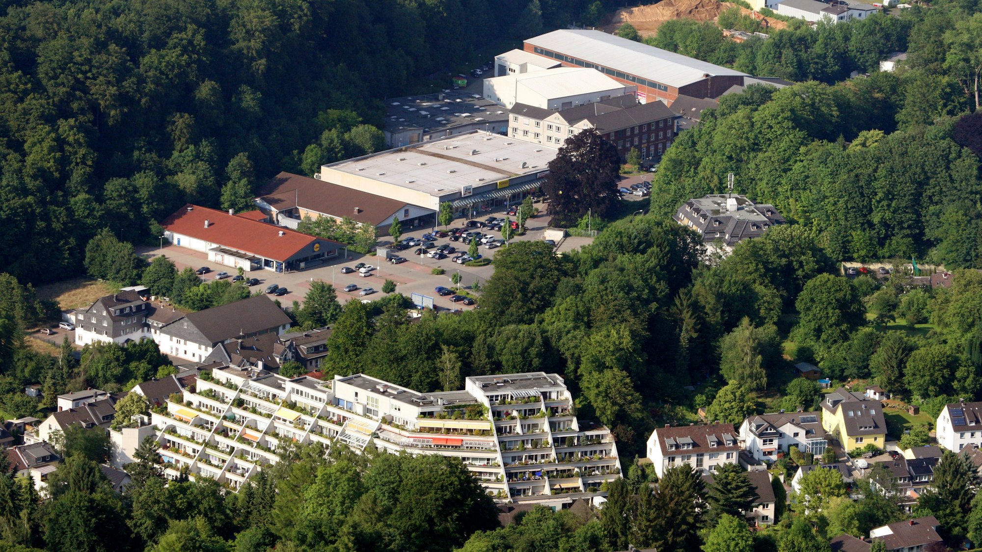 Eine Lufaufnahme mit einem terrassenartig angelegten Hochhaus, Einfamilienhäusern und einem großen Parkplatz vor einem Einkaufszentrum.