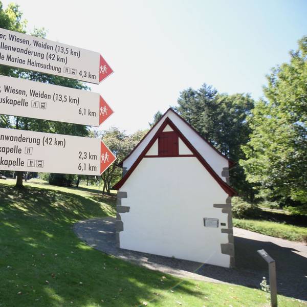 Schilder weisen den Weg für die Kapellenwanderung, im Hintergrund steht eine kleine Kapelle.