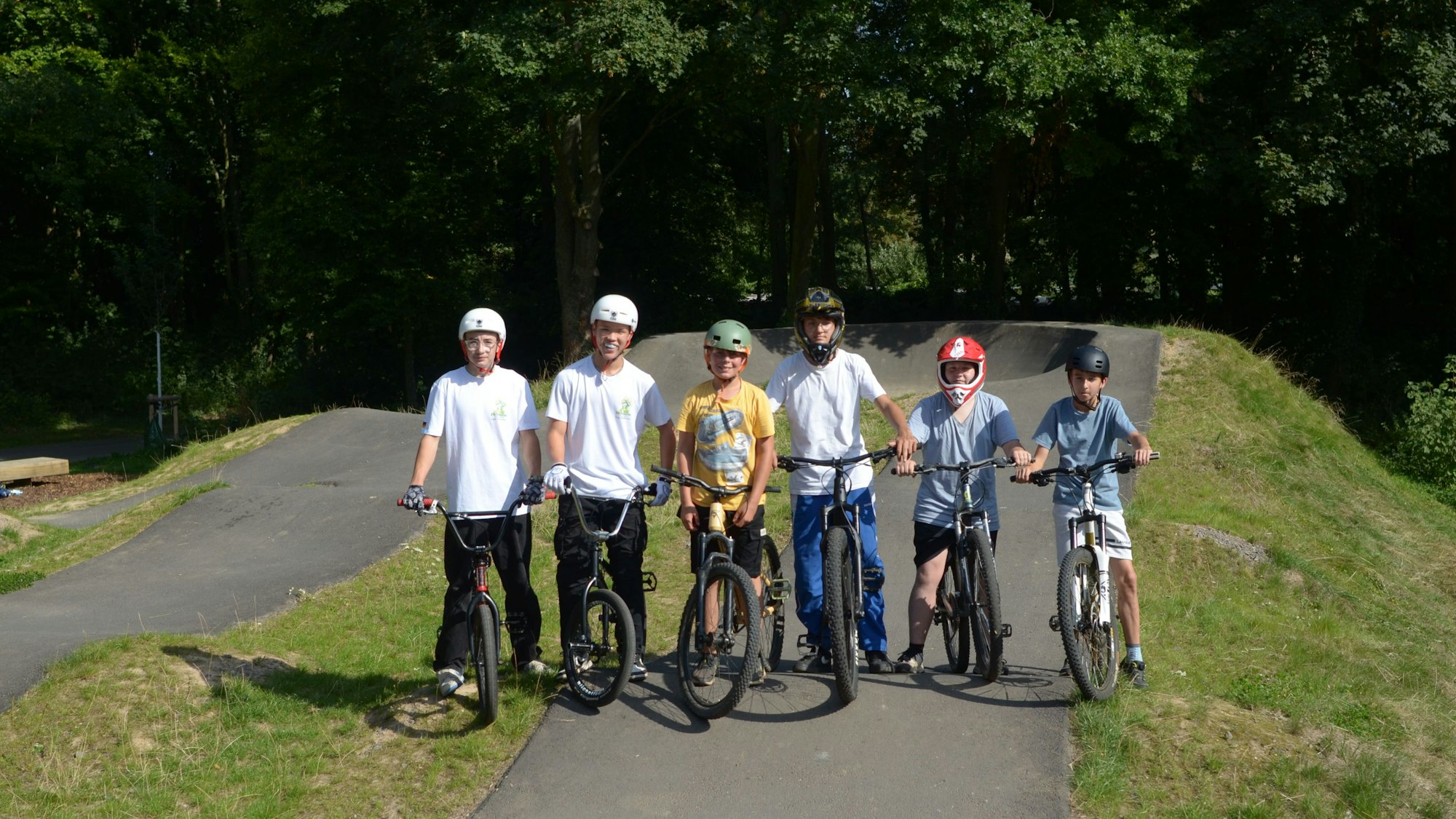 Sechs junge BMX-Fahrer, die Schutzhelme tragen, stehen mit ihren Rädern auf der asphaltierten Bahn.