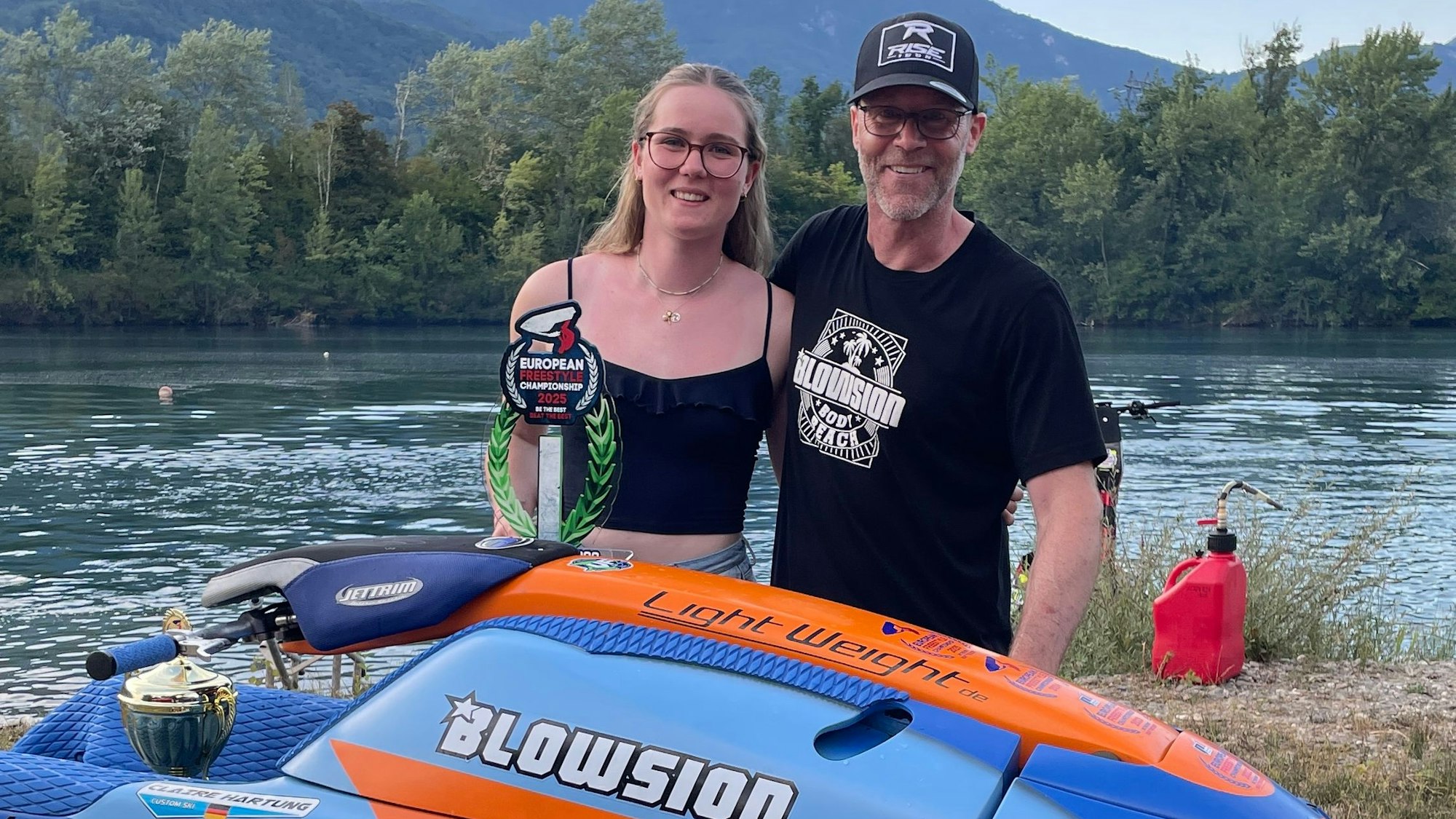 Eine junge Frau steht mit ihrem Vater hinter einem Jet-Ski und präsentiert ihren gewonnen Pokal.