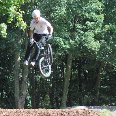 Das Bild zeigt einen BMX-Fahrer mit Schutzhelm, der aussieht, als stehe er nach einem Sprung neben seinem Fahrrad, dessen Lenker er hält, in der Luft.