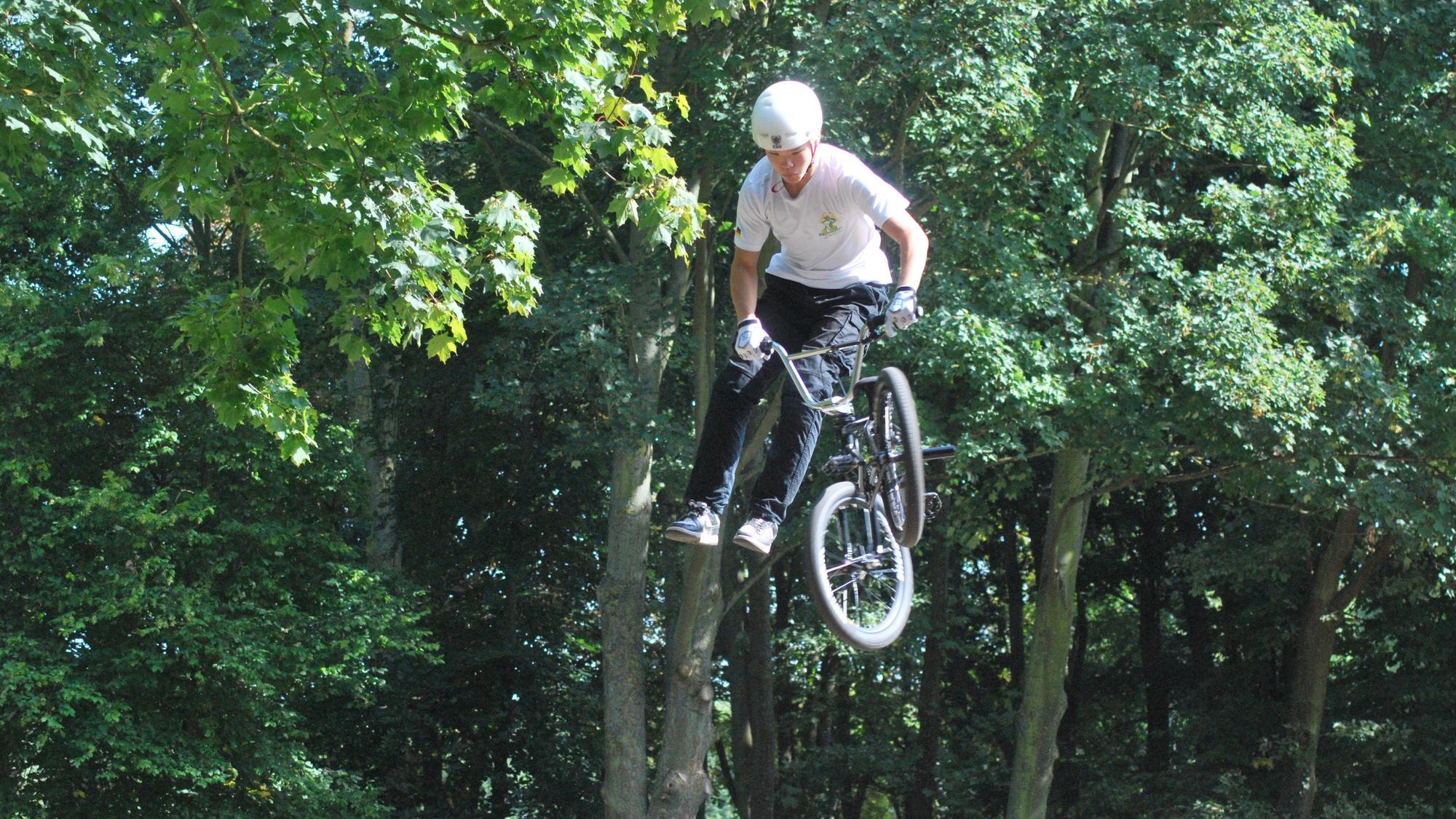 Das Bild zeigt einen BMX-Fahrer mit Schutzhelm, der aussieht, als stehe er nach einem Sprung neben seinem Fahrrad, dessen Lenker er hält, in der Luft.