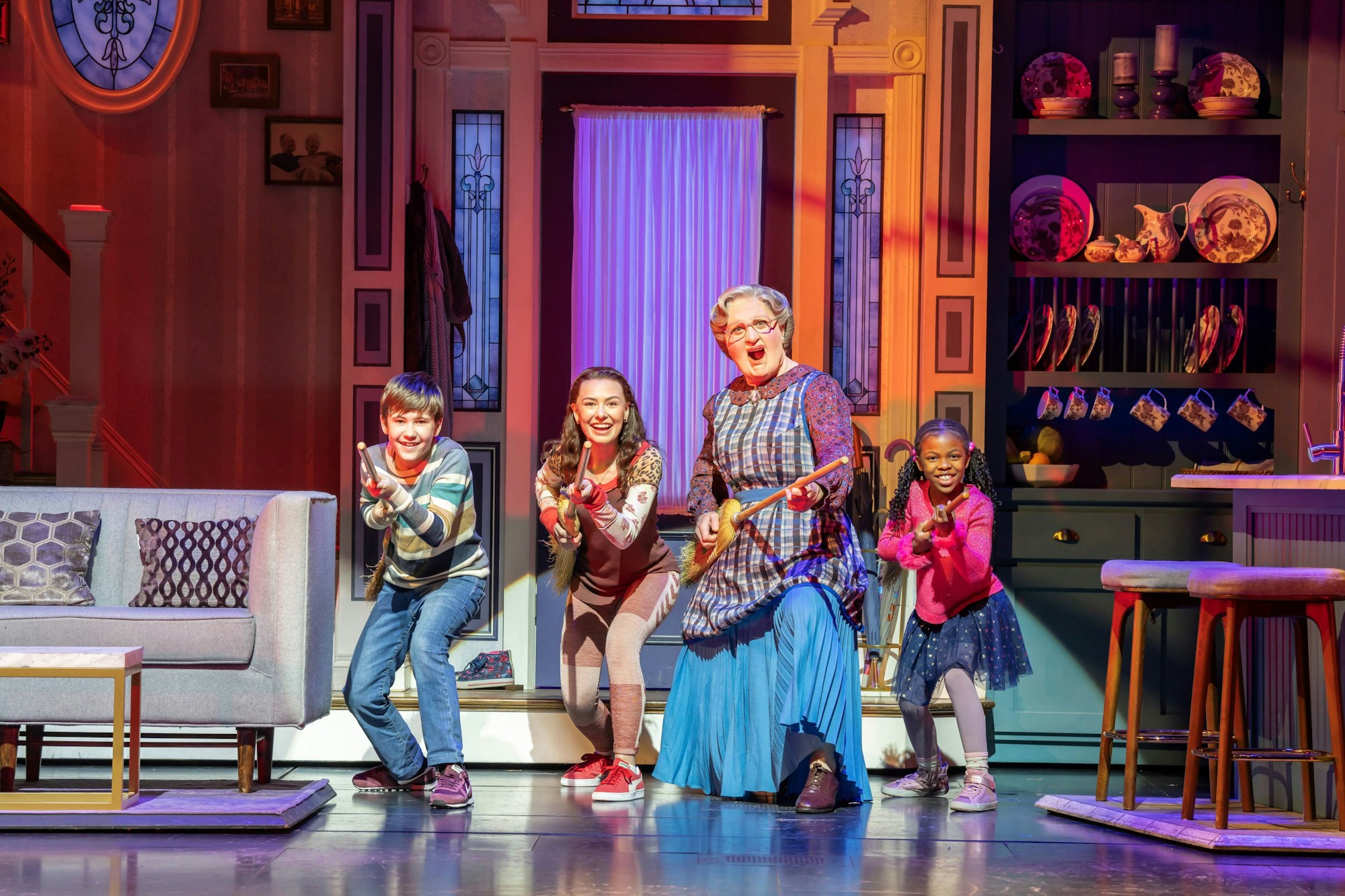 Musicalszene mit Mrs. Doubtfire und drei Kindern, die fröhlich mit Besen Gitarre spielen.