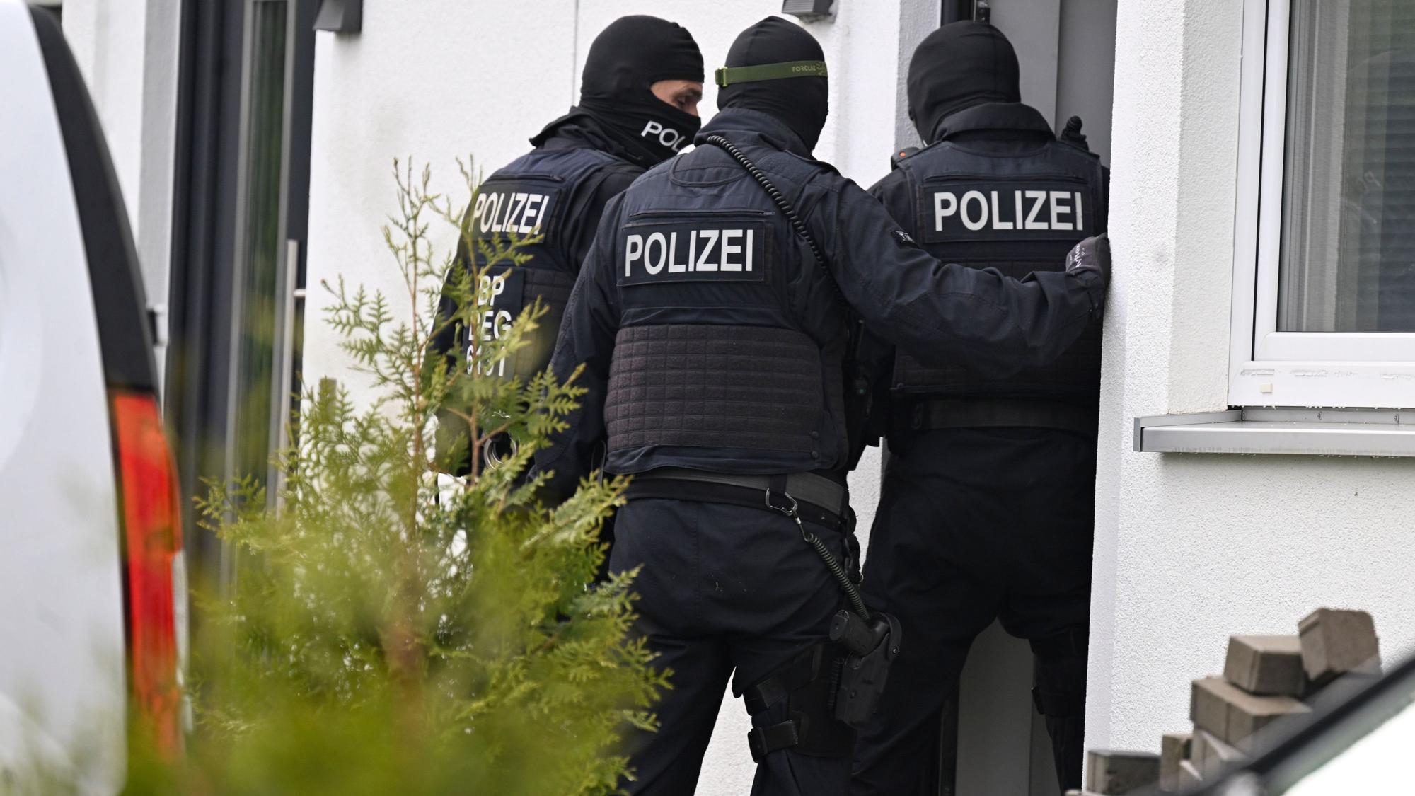 Drei schwarz vermummte Polizisten stehen vor dem Eingang eines weißen Wohnhauses.