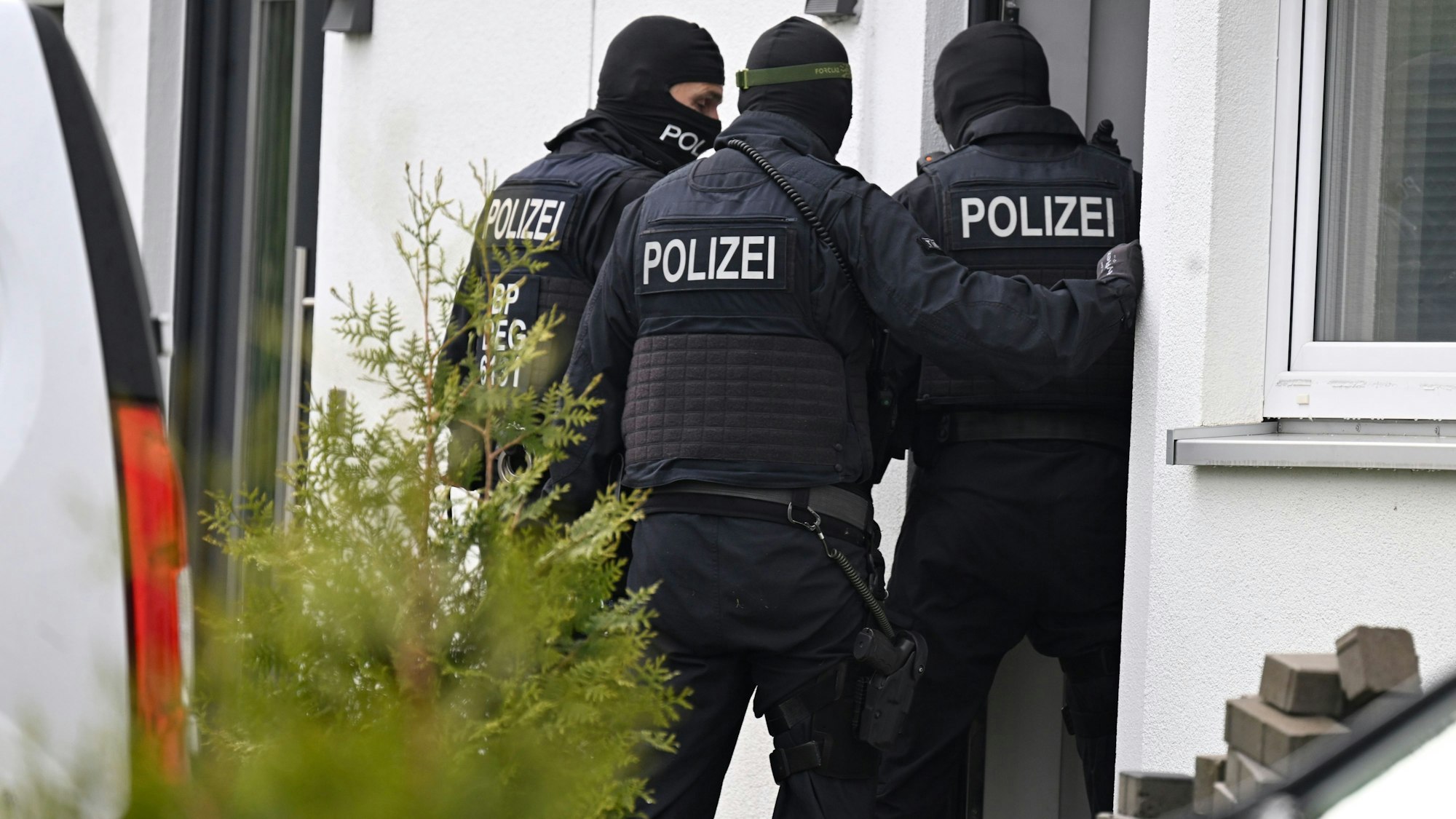 Drei schwarz vermummte Polizisten stehen vor dem Eingang eines weißen Wohnhauses.