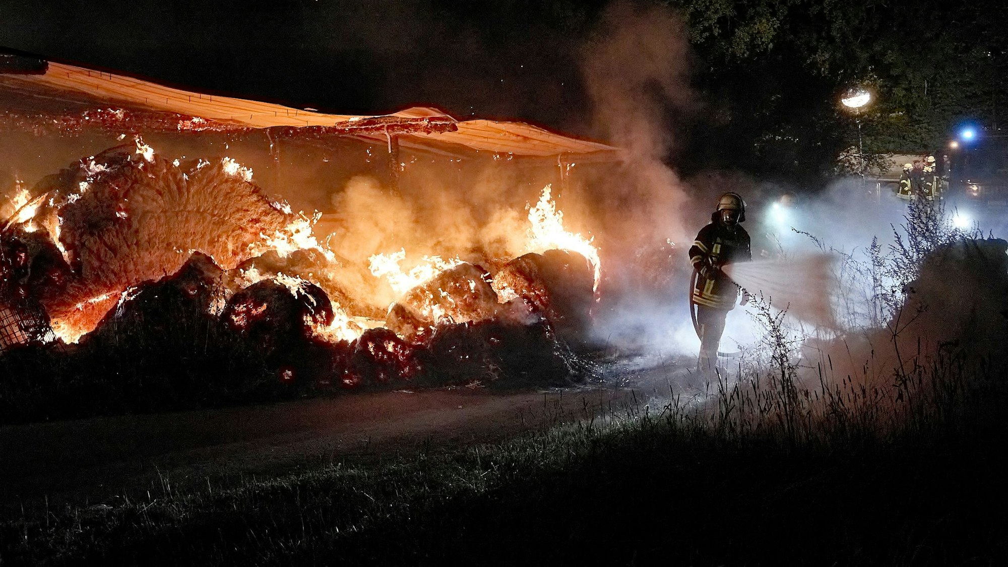 Vor dem Hintergrund brennende Strohballen hält ein Feuerwehrmann den Wasserstrahl auf das Gebüsch am rechten Bildrand.