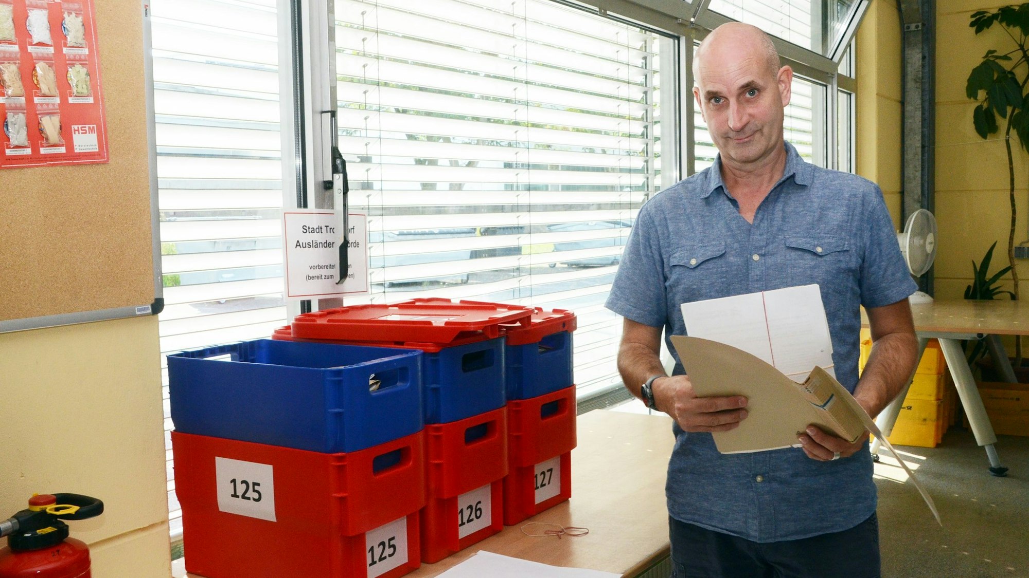 Ein Mann mit Glatze neben einem Tisch mit blauen und roten Kisten aus Kunststoff. In der Hand hält er einen Aktendeckel.