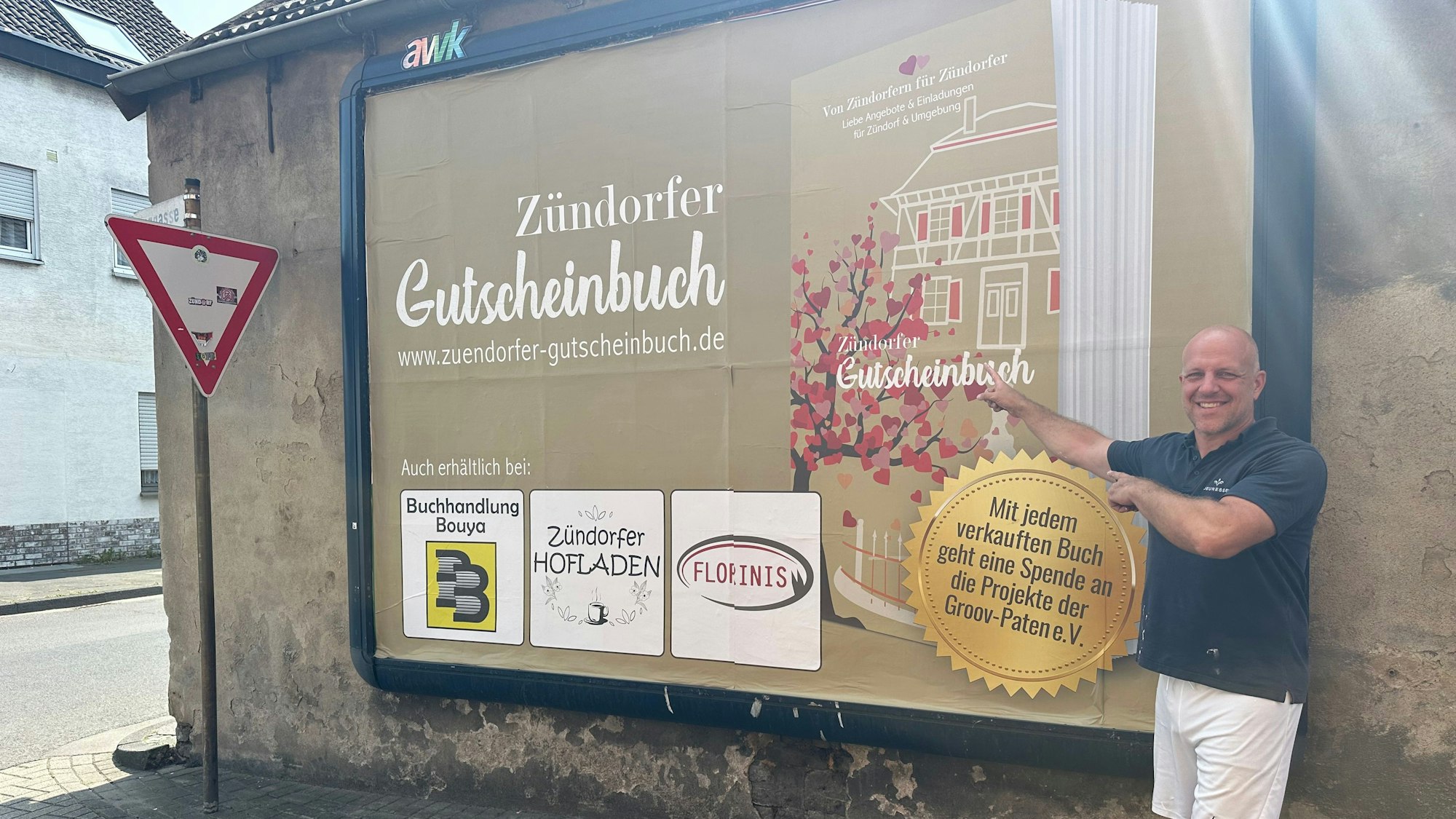 Ein Mann mit Glatze zeigt lächelnd auf ein Werbeplakat, dass das Zündorfer Gutscheinbuch zeigt.