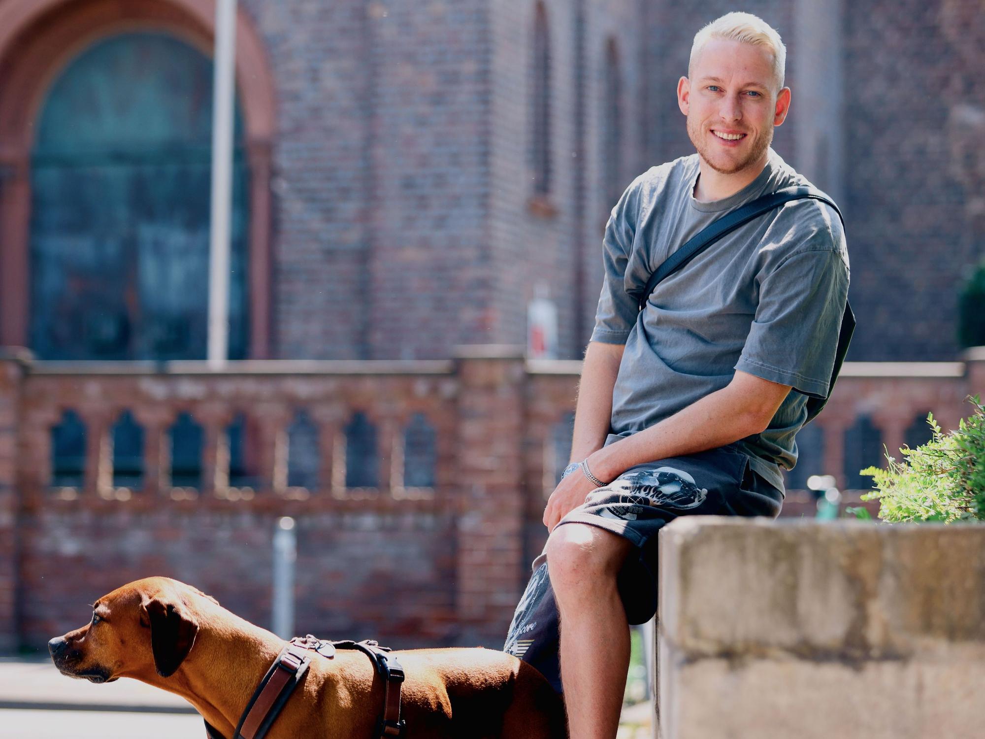 Ein junger Mann mit blondem kurzem Haar und einem braunen großen Hund sitzt auf dem Rand eines Blumenbeets, hinter ihm ist eine Kirche zu sehen.