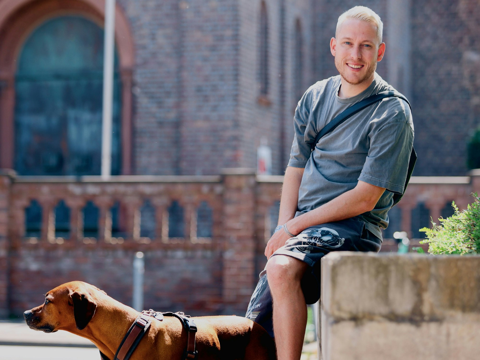 Ein junger Mann mit blondem kurzem Haar und einem braunen großen Hund sitzt auf dem Rand eines Blumenbeets, hinter ihm ist eine Kirche zu sehen.