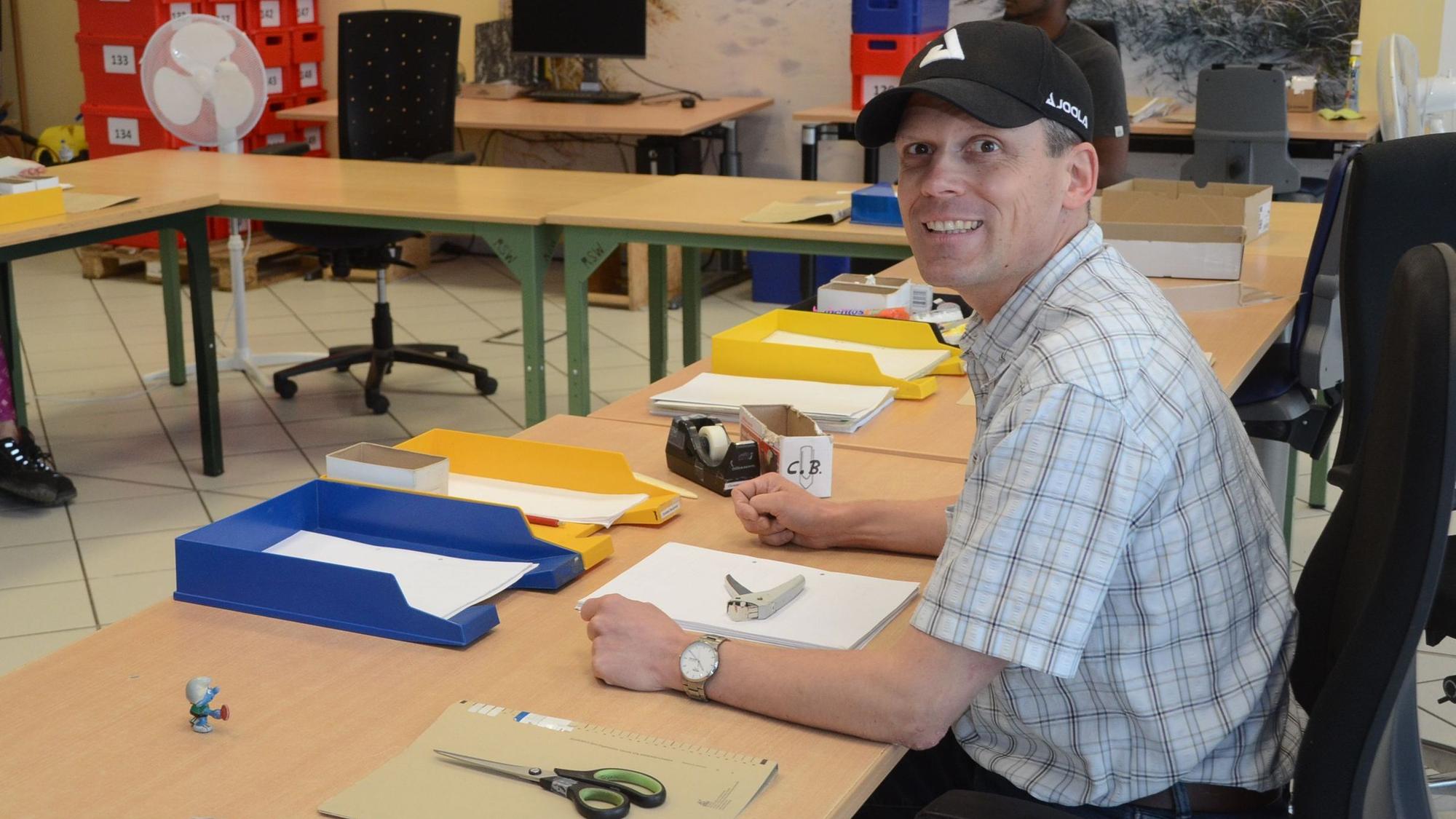 Ein Mann mit schwarzer Baseballkappe an einem Arbeitstisch mit Büromaterialien.
