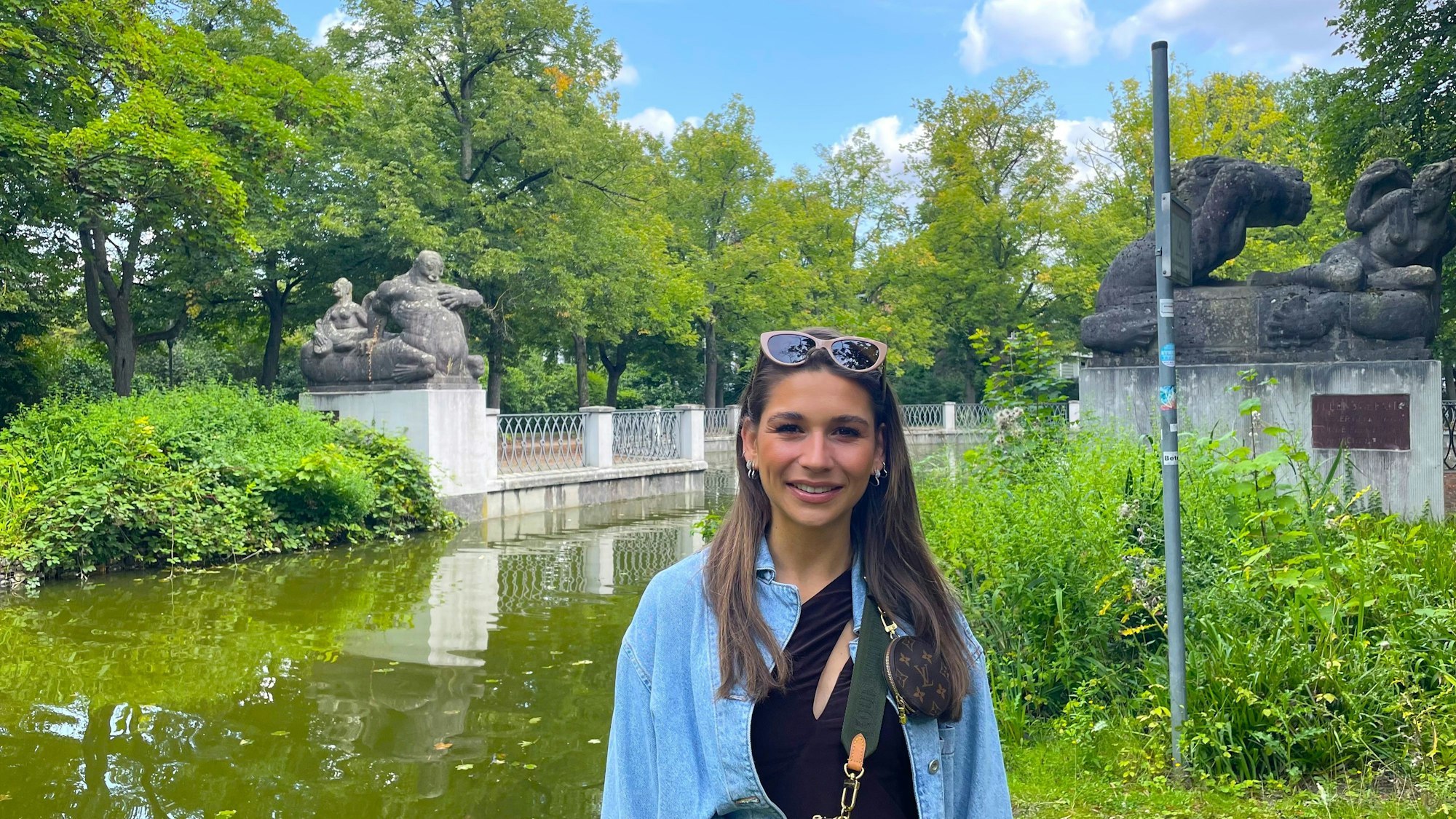 Eine junge Frau mit langem Haar steht vor einem Kanal, an dessen Ufern zwei Steinskulpturen stehen.