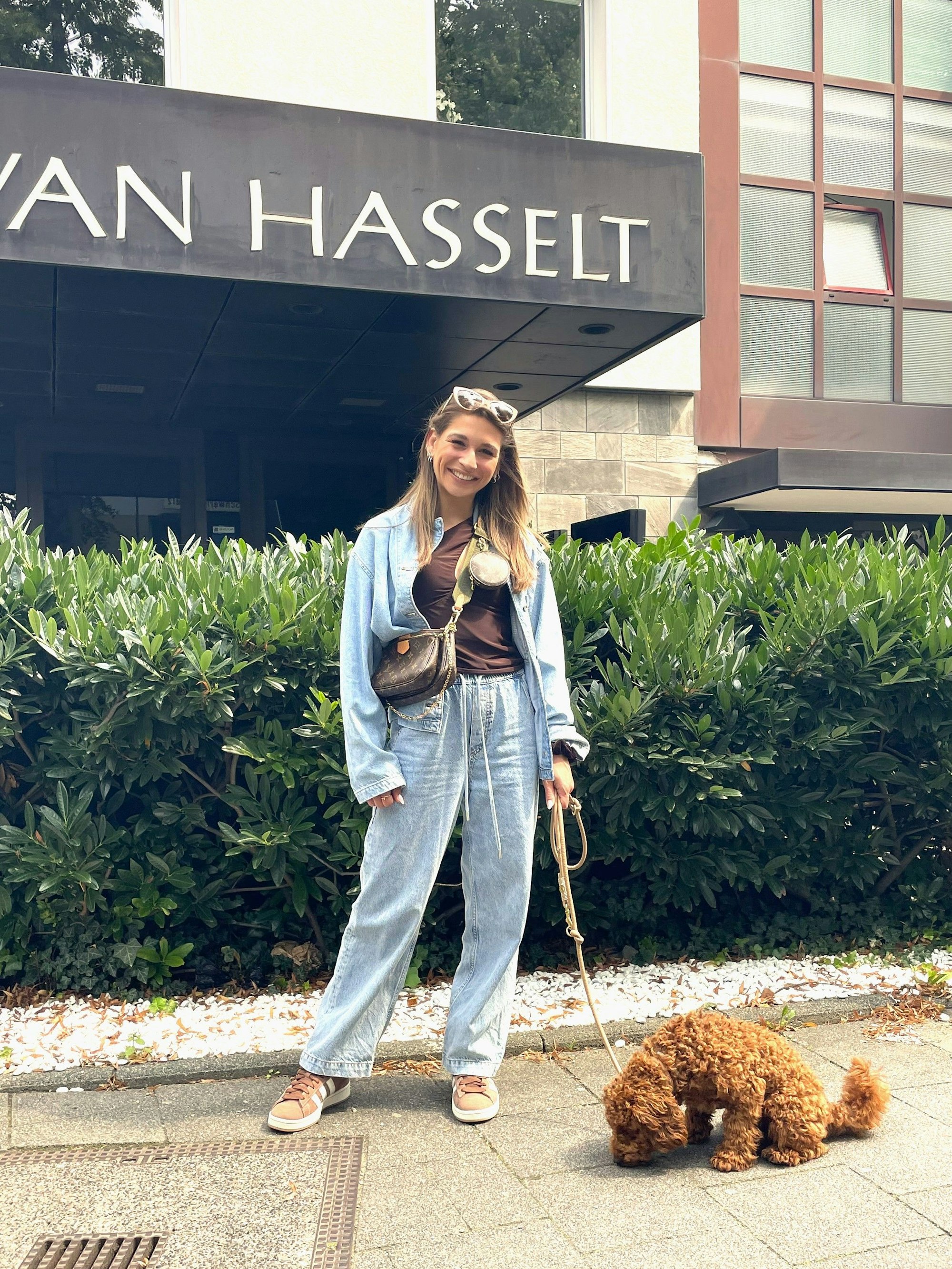 Eine junge Frau steht vor einer schulterhohen Hecke, an der Lein einen braunen Zwergpudel.