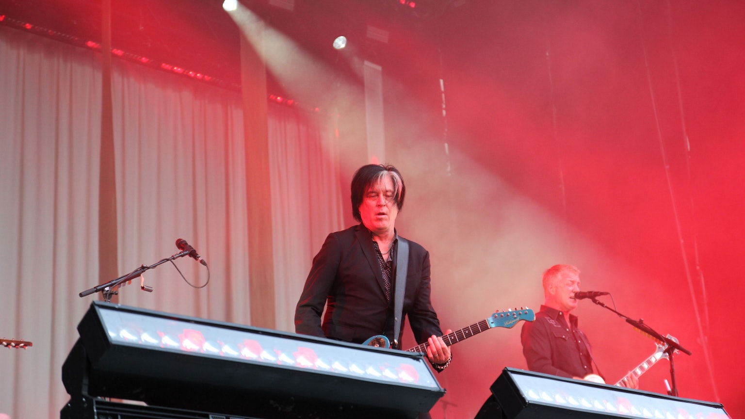 Zwei Gitarristen stehen auf einer in rotem Licht getauchten Bühne. Es sind Troy Van Leeuwen und Josh Homme von Queens Of The Stone Age.