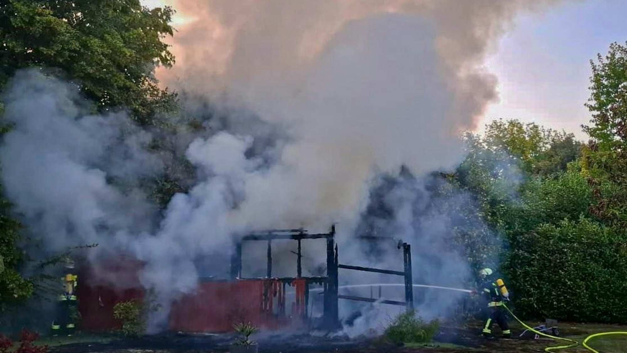 Weiße Rauchschwaden über einem abgebrannten Gartenhaus. An der rechten Seite eine Feuerwehreinsatzkraft in voller Montur und Schlauch.
