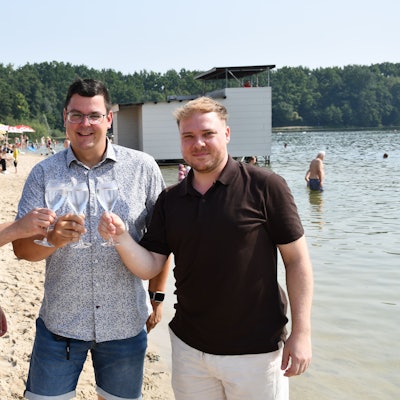 Das Foto zeigt drei Männer lockerer Kleidung im Strandbad, die mit Sektgläsern anstoßen.