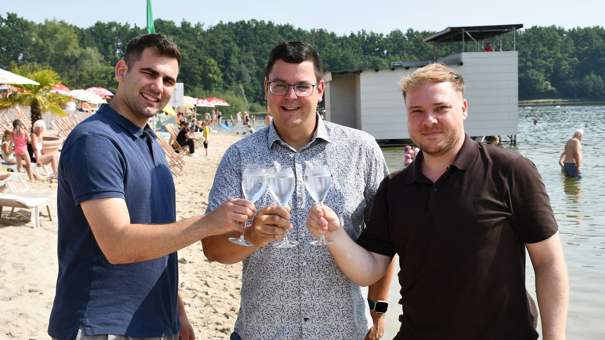 Das Foto zeigt drei Männer lockerer Kleidung im Strandbad, die mit Sektgläsern anstoßen.