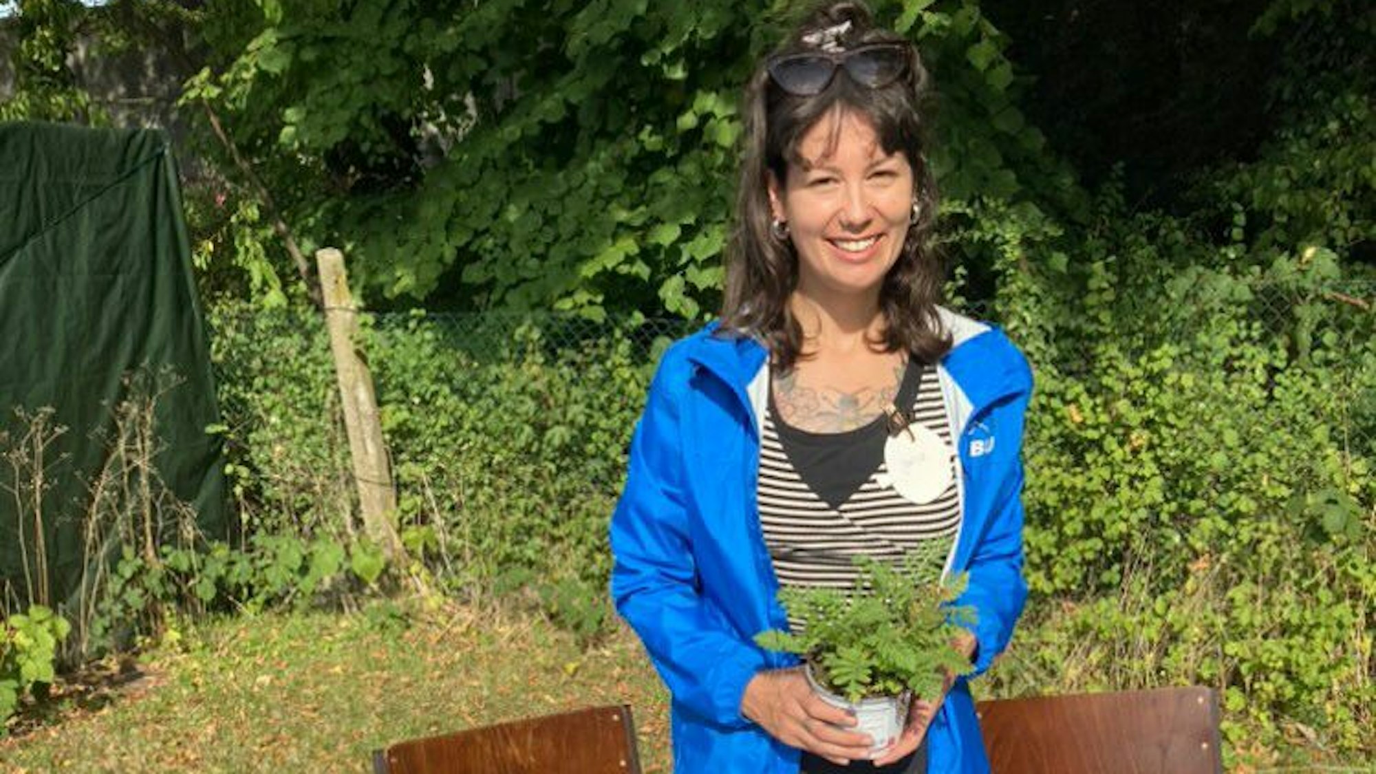Auf dem Bild trägt Jana Romero eine blaue Trainingsjacke. Sie hält einen Topf mit einer Pflanze. Im Hintergrund jede Menge Grün.