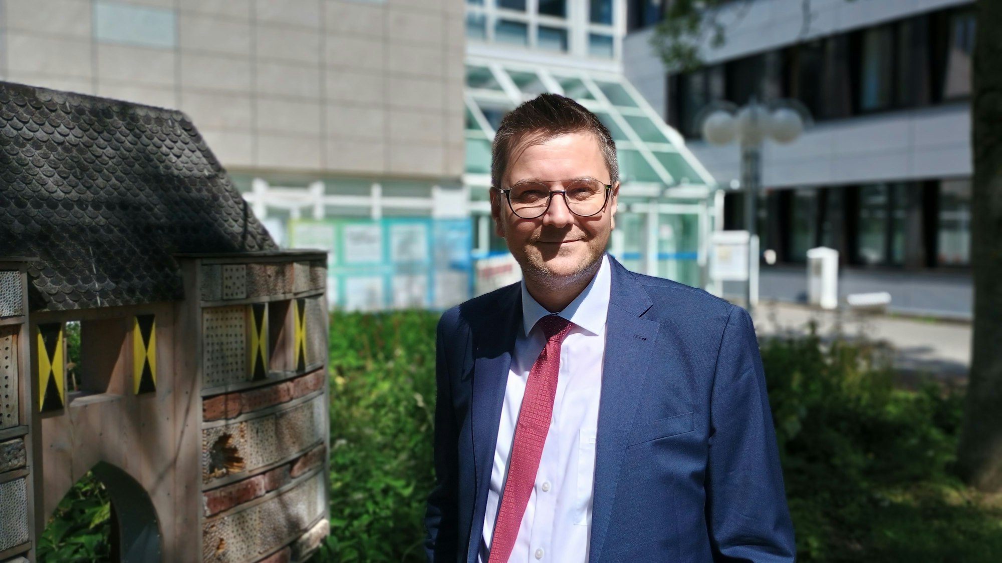 Rekewitz trägt einen dunkelblauen Anzug und eine rote Krawatte. Er steht vor dem Rathaus der Stadt Bergheim, neben sich ein Insektenhoel in Form des Aachener Tors.