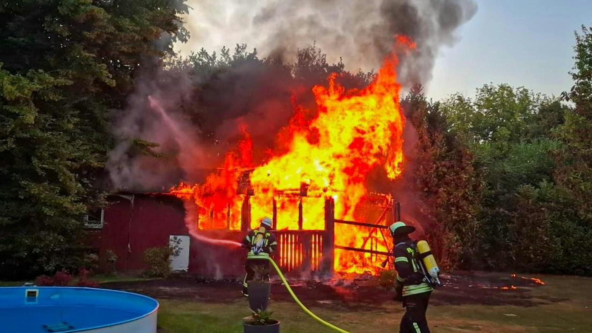 Ein Gartenhaus brennt lichterloh. Im Vordergrund Feuerwehrleute im Einsatz.