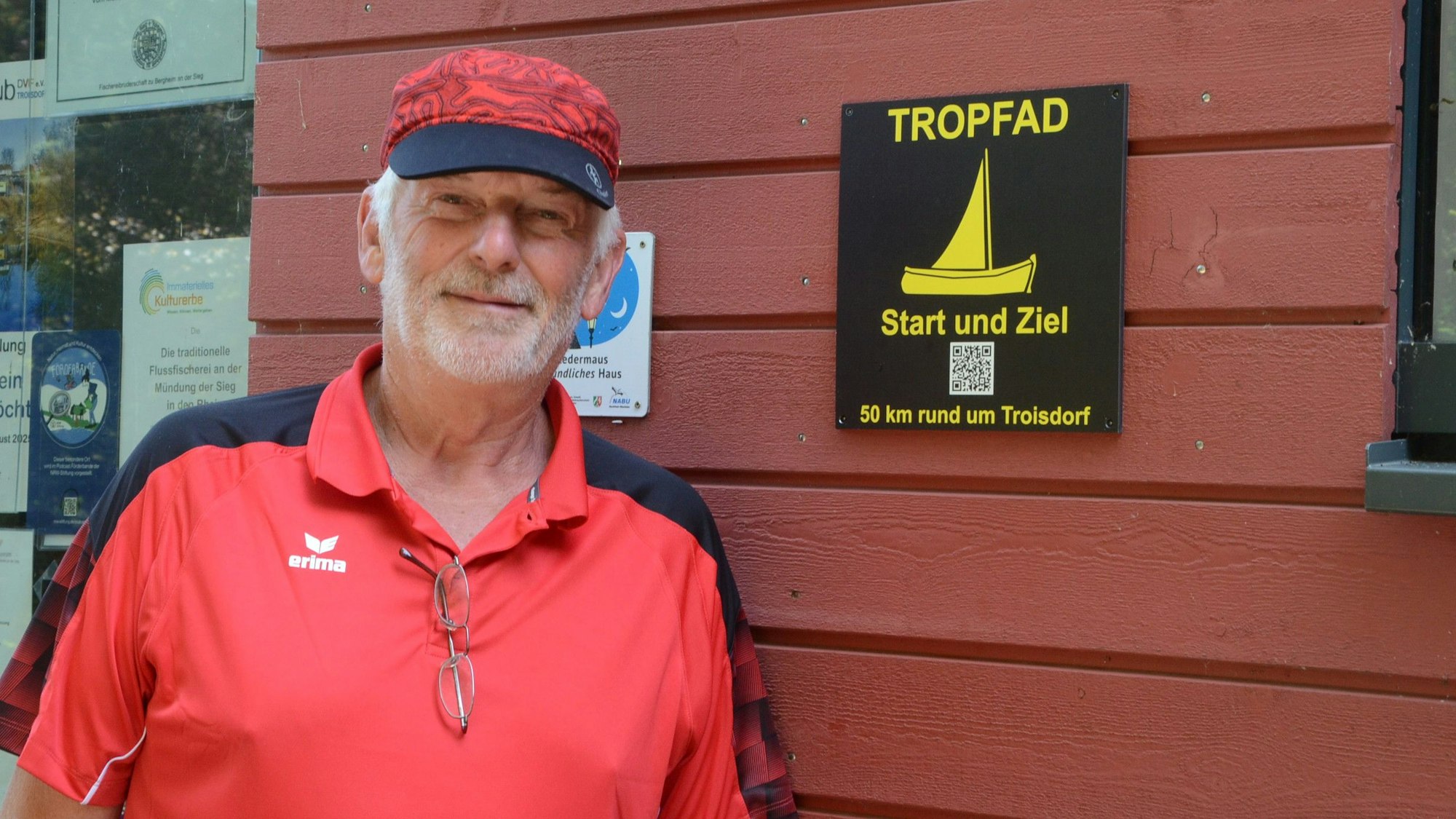 Ein Mann im roten Hemd und mit einer roten Kappe steht vor einer roten Holzwand. Auf einem schwarzen Schild steht in gelber Schrift Tropfad Start und Ziel.50 km rund um Troisdorf