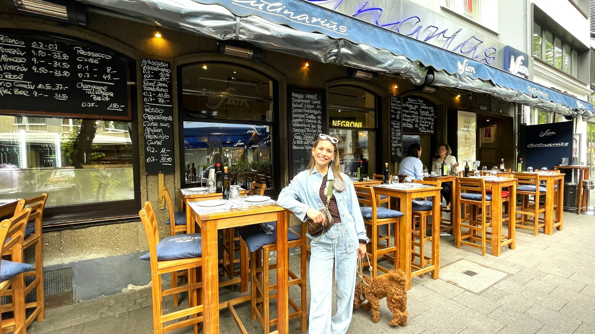Eine junge Frau mit Sonnenbrille auf dem Kopf steht an einem Stehtisch vor einem italienischen Restaurant.