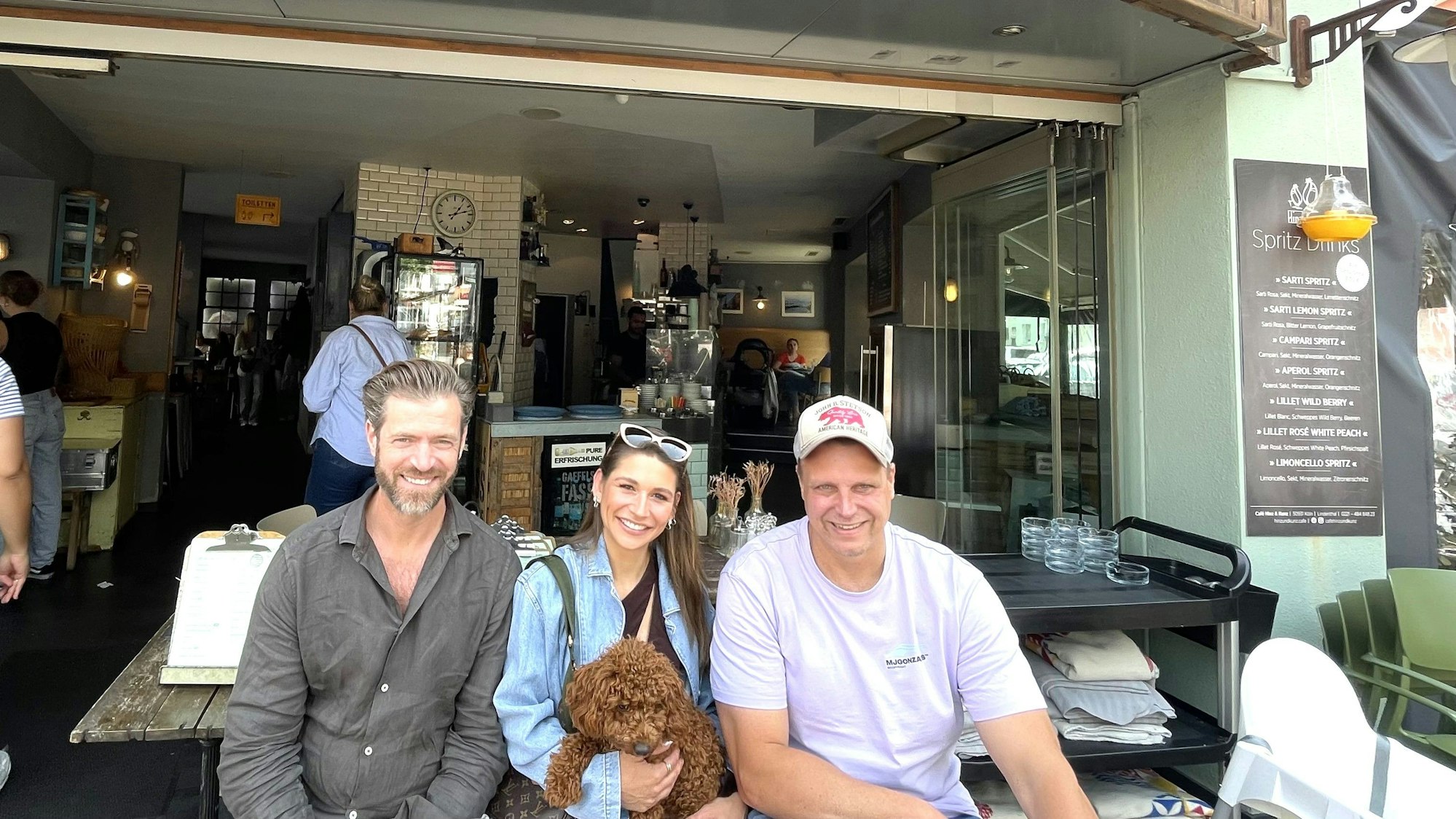 Eine junge Frau mit einem Zwergpudel auf dem Schoß sitzt vor einem Café, links und rechts von ihr je ein Mann, es sind die Inhaber des Cafés Hinz und Kunz.