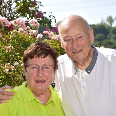 Das Paar steht vor einem Busch in voller Blühte. Anna Schiffer trägt ein grünes Hemd, Bruno Schiffer ein weißes.