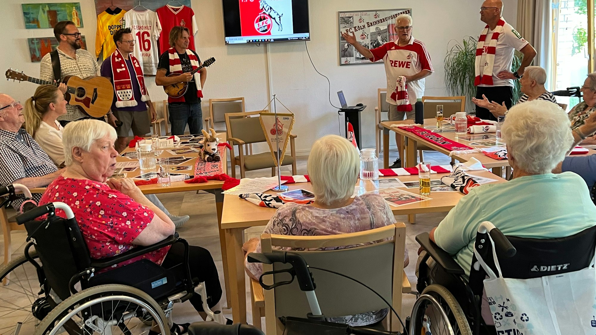Drei Musiker und zwei Männer in FC-Trikots stehen in einem Raum, in dem ältere Menschen teils in Rollstühlen sitzen.