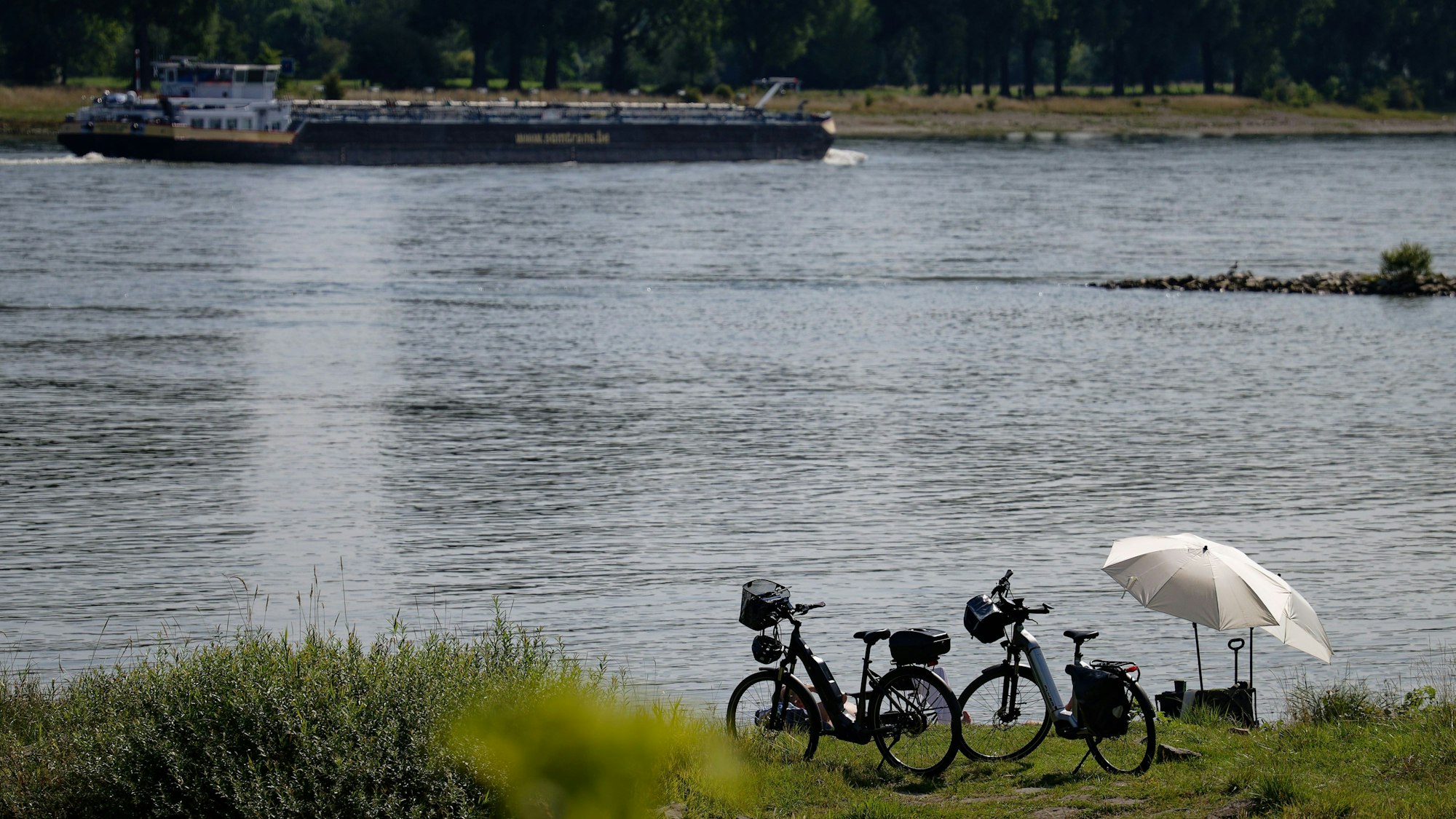 Fahrräder und Sonnenschirme stehen am Rheinufer bei Köln-Langel. Im Hintergrund ein Frachtschiff.