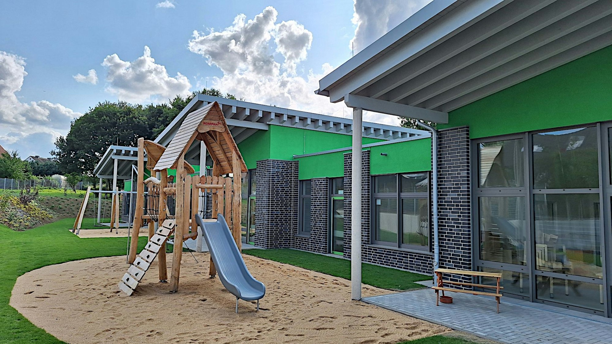 Vor den Gebäuden der neuen Kita, die Pultdächer haben steht ein Klettergerüst mit Rutsche.