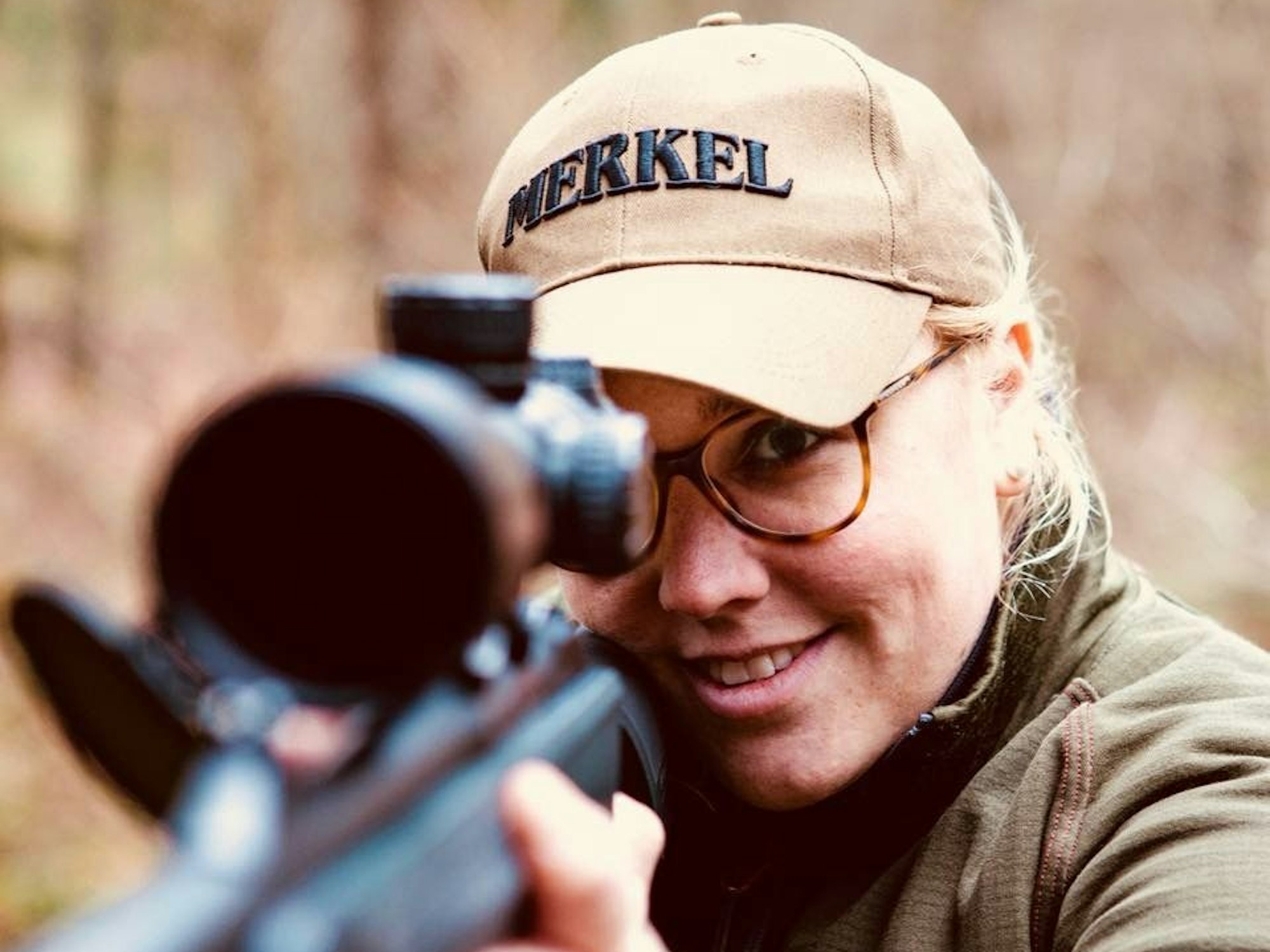 Die Jägerin trägt eine Mütze und eine Brille. Sie schaut durch das Zielfernrohr einer Waffe.