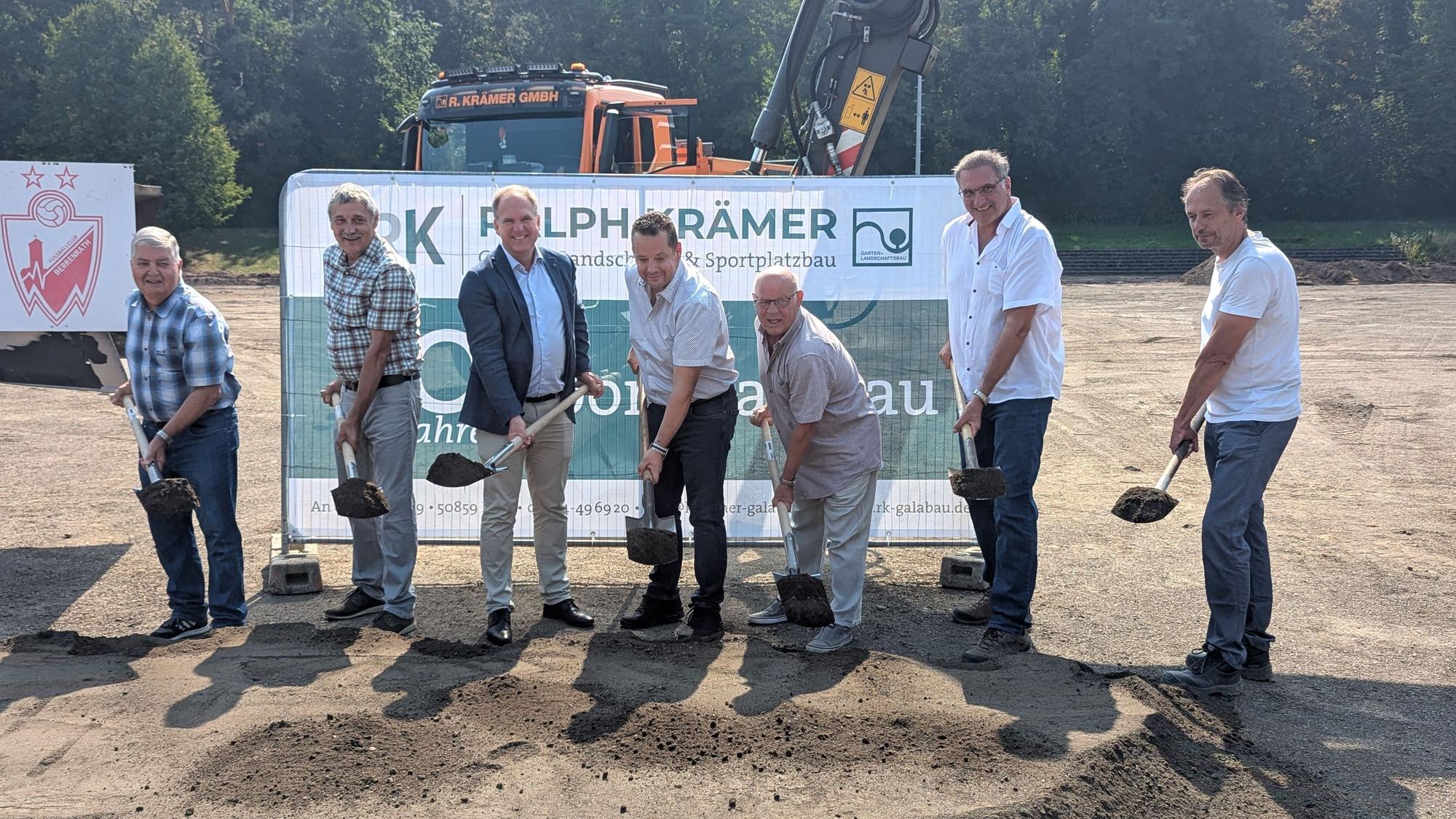 Das Foto zeigt sieben Männer mit Spaten vor einem Banner und Baumaschinen auf dem Sportplatz.