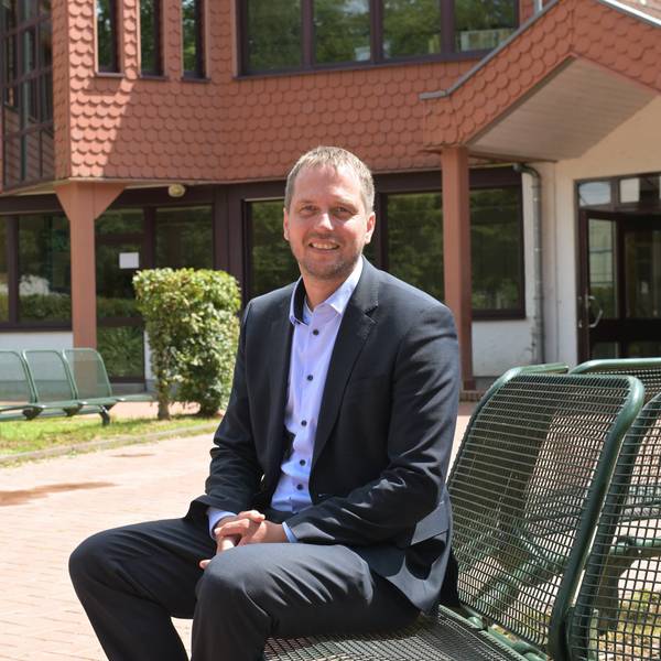 Ein Mann sitzt in einem dunklen Anzug vor einer modernen Grundschule.