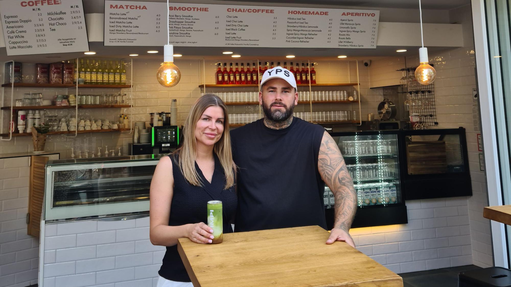Eine Frau mit blondem Haar steht neben einem Mann mit Bart und Basecap hinter dem Stehtisch eines Cafés.
