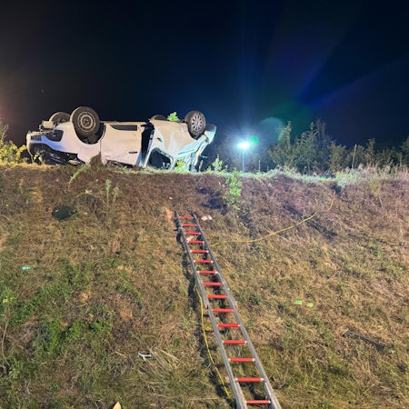 In der Mitte des Bildes ein Fahrzeug auf dem Dach liegend auf einer Böschung. Eine Feuerwehrleiter führt zum Fahrzeug. Links mehrere Einsatzkräfte.