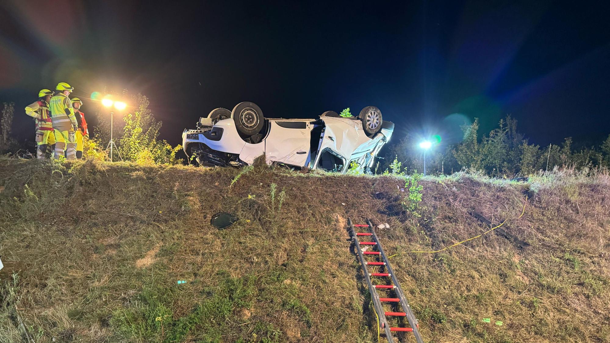 In der Mitte des Bildes ein Fahrzeug auf dem Dach liegend auf einer Böschung. Eine Feuerwehrleiter führt zum Fahrzeug. Links mehrere Einsatzkräfte.