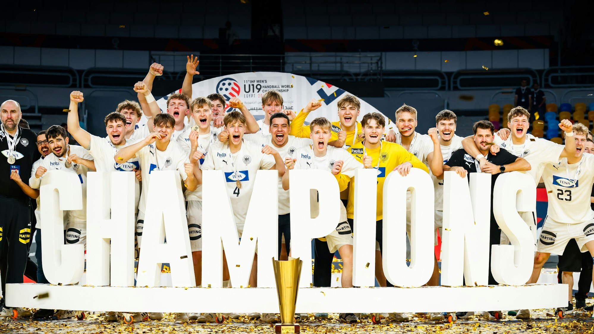 Die U19-Handballer stehen hinter dem Weltmeisterpokal und dem Schriftzug Champions und jubeln.