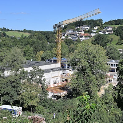 Ein großer Kran steht über unfertigen Rohbauten in einem grünen Tal.