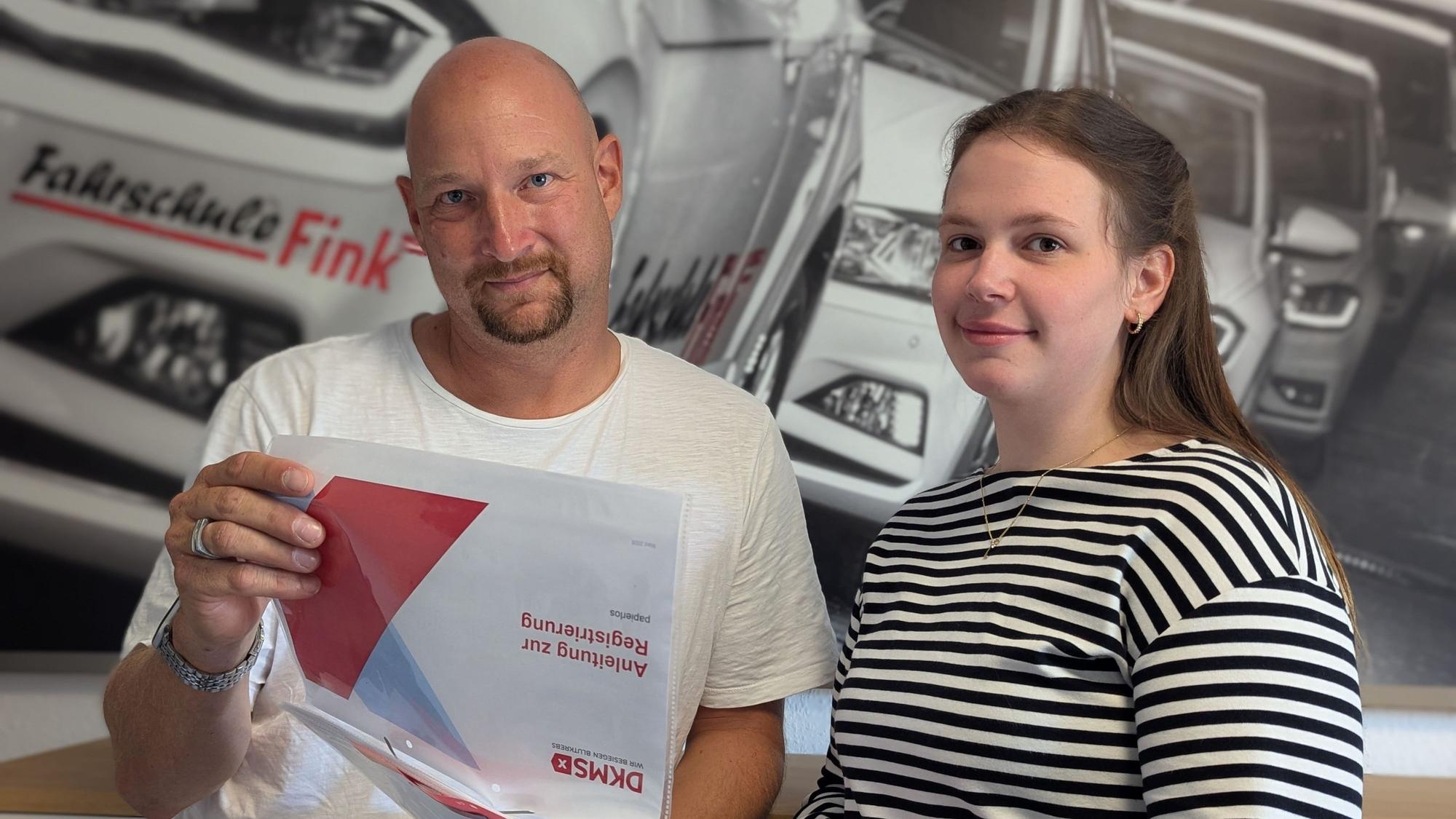 Das Foto zeigt zwei Personen mit einer Infomappe des DKMS vor einem Foto in der Fahrschule Fink in Hürth.